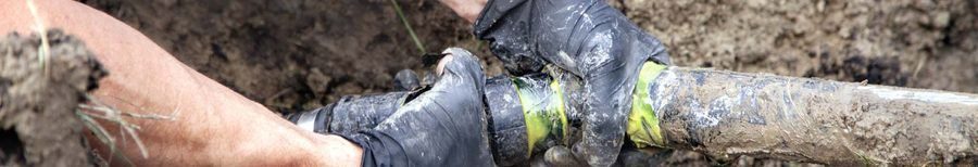 Close-up of gloved hands gripping a muddy pipe joint outdoors, indicating plumbing or trench work.